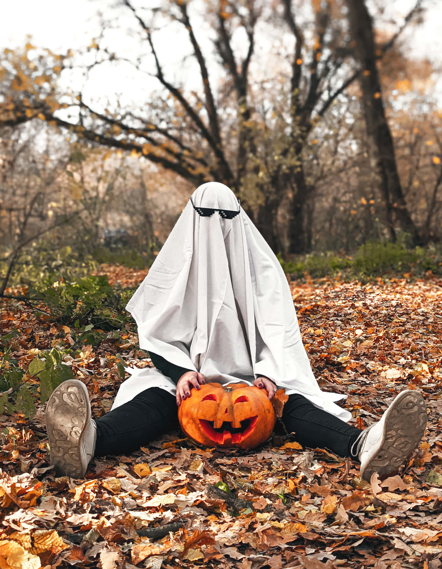 Person in ghost costume with glasses and pumpkin sitting down in forest